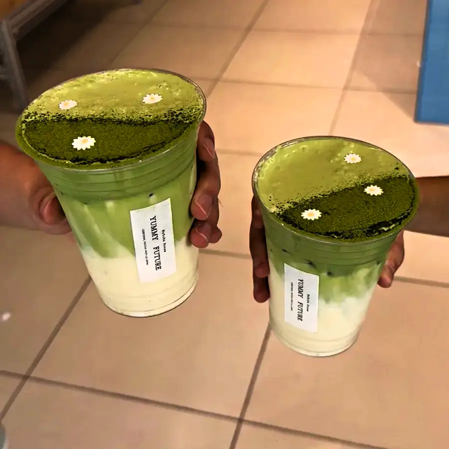 Two matcha lattes held up inside the shop, tables in the background